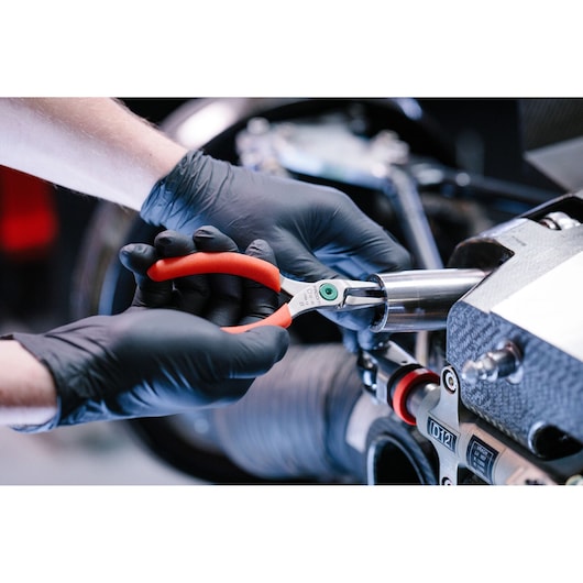 A person wearing black gloves uses red-handled Facom pliers to work on a metallic mechanical component.