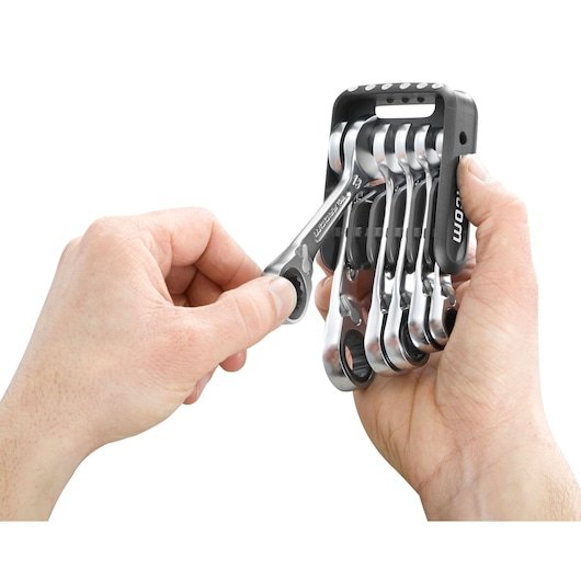 Two hands holding a set of Facom ratcheting combination wrenches organized in a black plastic holder, with one wrench being removed.