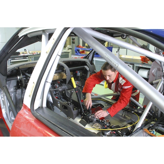 Facom A_442, mechanic in red suit working on race car interior, roll cage and car parts visible.