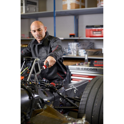 Facom ATPA4V.J12APB mechanic using hand tool on race car in workshop, red tool storage and shelves in background.