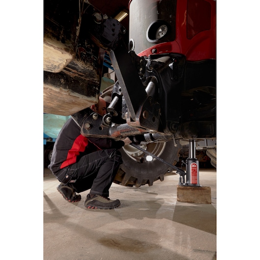 Photo of Facom DL12BTI hydraulic bottle jack lifting large machinery indoors, mechanic in work clothes adjusting under vehicle.