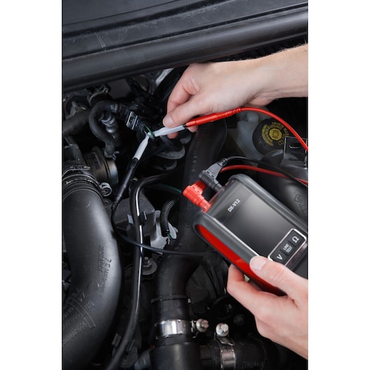 Photo of Facom DX_SET automotive tester being used on engine wires, close-up of hands and device in action.