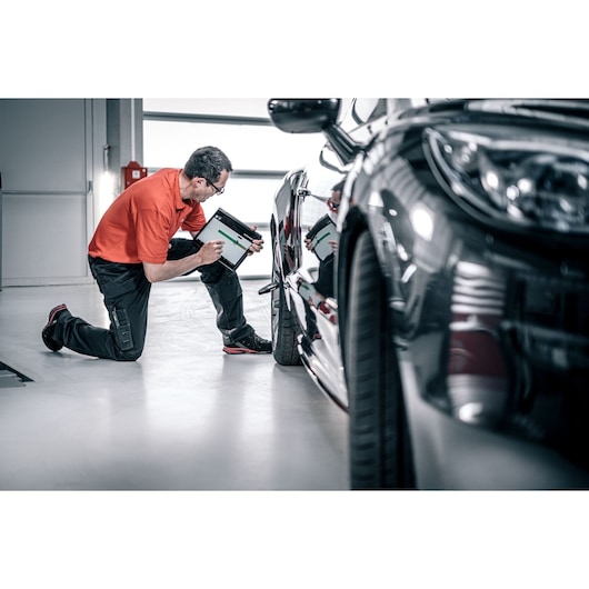 Facom DX_TSCANPB automotive technician kneeling, scanning car body with tablet in workshop.
