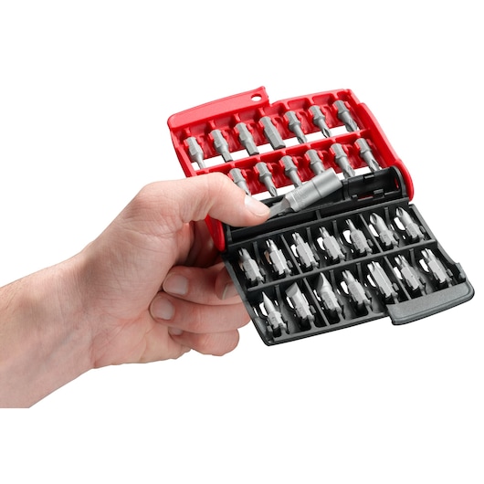 A hand holding an open plastic case containing a variety of screwdriver bits organized in rows. The case is red and black, and several metal bits are visible.