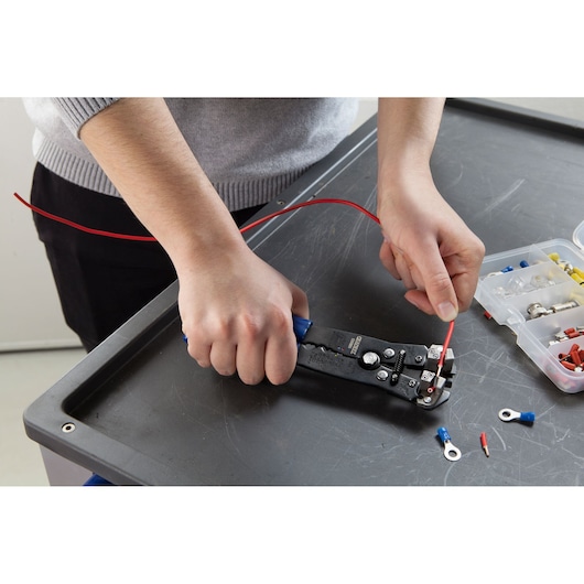 Facom E050105 wire stripper tool in use on a red wire, with electrical connectors nearby on a worktable.