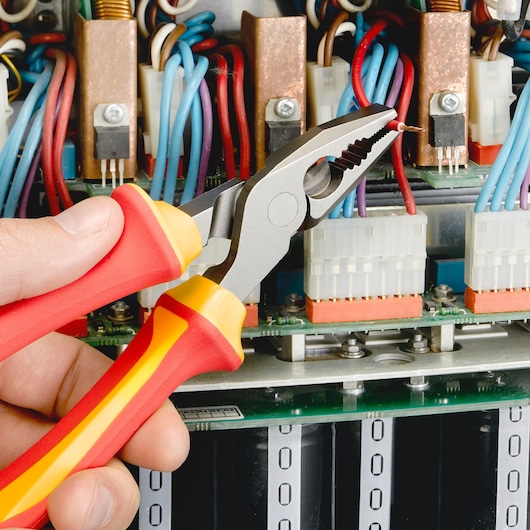 Facom E050412 insulated combination pliers held by hand, gripping wire near electrical circuit.