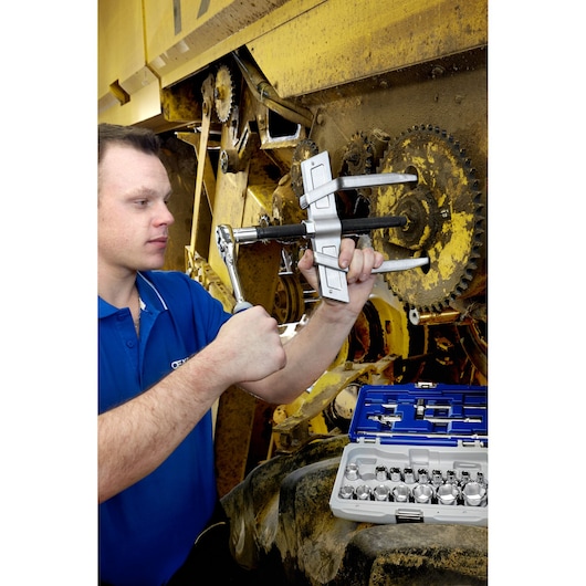 Facom E096005 gear puller tool in use on industrial machinery, mechanic holding ratchet, socket set visible.