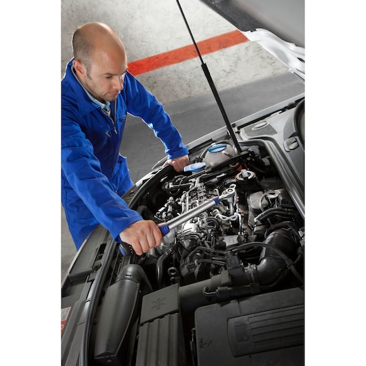 Facom E100105 torque wrench used by mechanic in blue coverall on car engine, view from angle above.