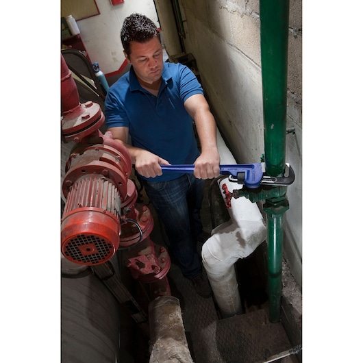 Facom E117824 pipe wrench in use on green pipe, blue handle visible, indoor industrial setting.
