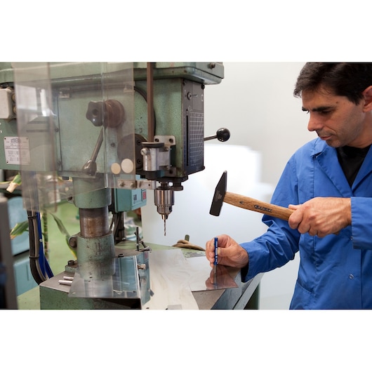 Facom E418236 machinist marking metal sheet with hammer and punch near industrial drill press.