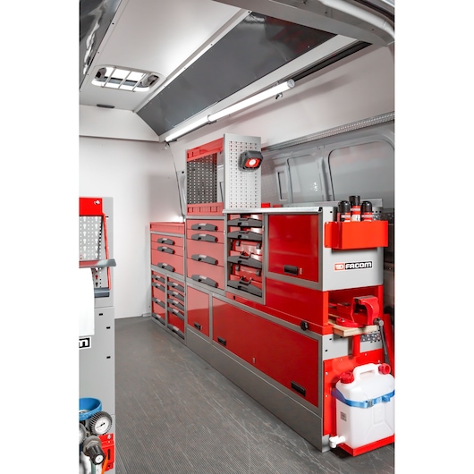 Interior of a service van equipped with Facom red tool storage units and drawers, organized along the side wall. The setup includes multiple drawers, shelves, and compartments, with tools and accessories arranged neatly, and branding visible on the storage. A white water container and various maintenance products are also present.