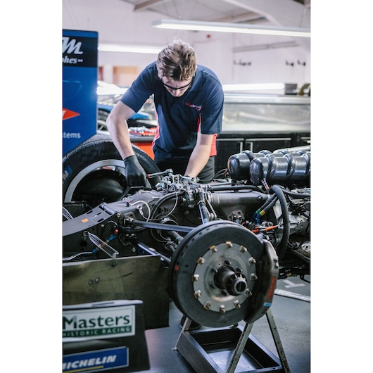 A mechanic working on the exposed engine and braking system of a racing car in a garage. The mechanic is wearing gloves and a dark polo shirt, and is using tools to adjust components of the car.