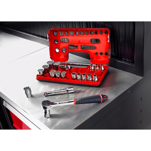 A red Facom socket and ratchet set is displayed open on a metal workbench, showing multiple sockets, extensions, and a ratchet tool. A few pieces are placed nearby on the bench.