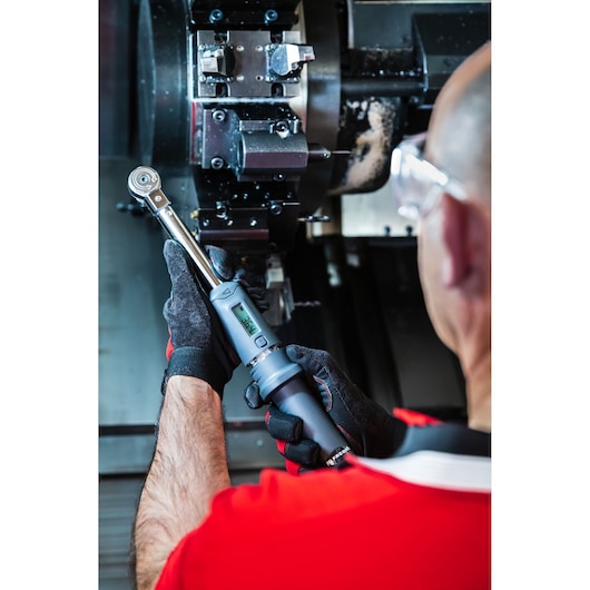 Facom S.307A100 digital torque wrench held by worker with gloves near machinery, industrial setting.