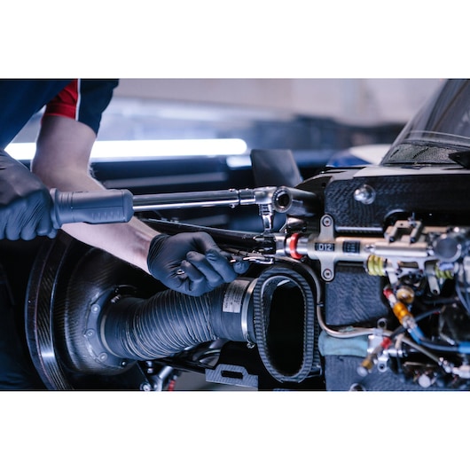 A mechanic wearing black gloves uses a Facom ratchet and socket tool to work on a complex part of a car engine, with visible cables and components.