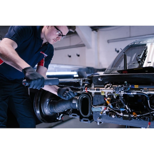 A mechanic wearing black gloves uses a Facom ratchet wrench to work on the exposed inner components of a car in a workshop.