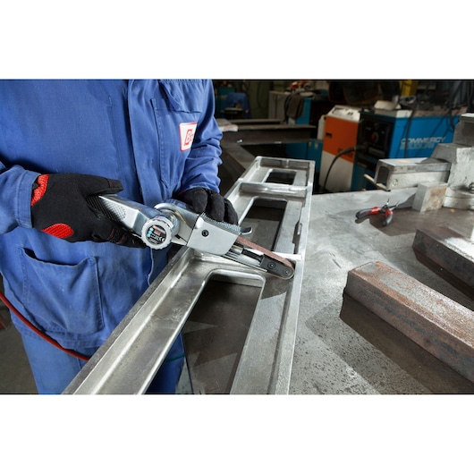 Photo of a worker using a Facom V_403F belt sander tool on a metal frame in an industrial workshop.